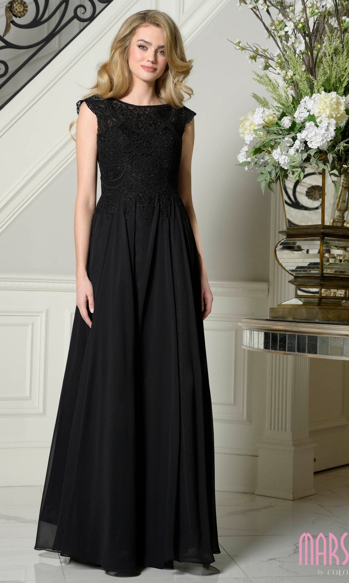 A woman with blonde hair wears the Marsoni M238, a black long formal dress, standing indoors near a staircase and floral arrangement.